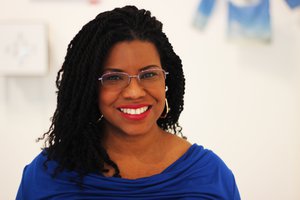 A headshot of a smiling woman with dark hair, glasses, and wearing a blue top.