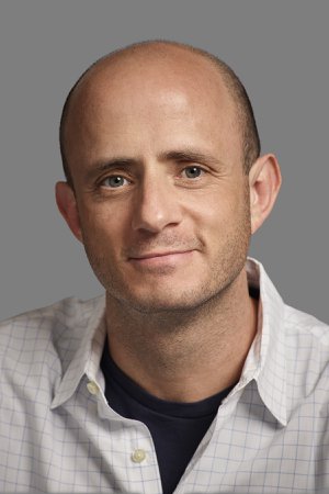 A headshot of a smiling, balding man in a checkered white dress shirt.