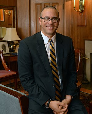 A smiling man in a suit and glasses seated in a chair in an office or study.