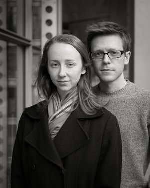 A black and white photo of a young woman standing in front of a young man in glasses.