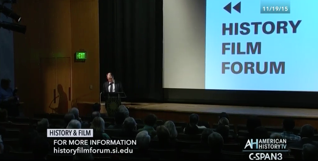 A man speaking at a lectern with the History Film Forum displayed on a large screen behind him